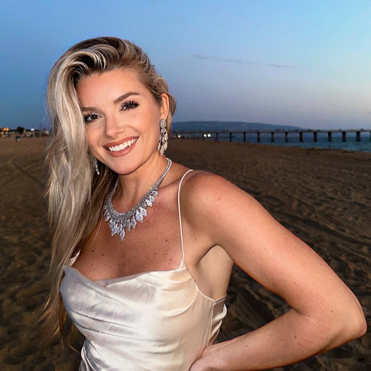 A woman standing on a beach at sunset wearing a white dress and the Amira necklace.